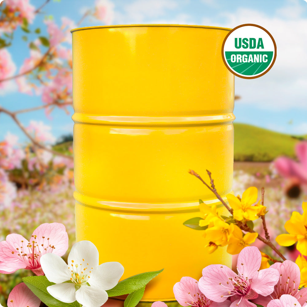 USDA Organic bulk honey drum surrounded by colorful spring blossoms in a sunny natural landscape.